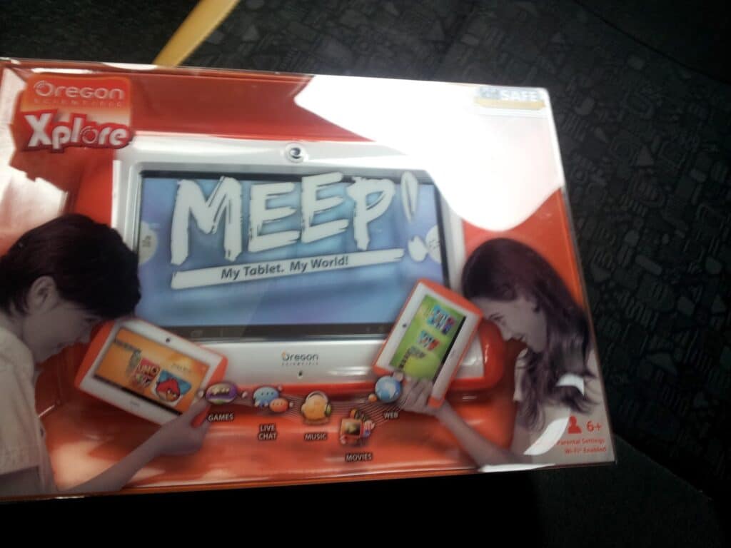 A boxed Oregon Scientific Xplore Meep! kids’ tablet is shown. The packaging features a large image of the tablet with “MEEP! My Tablet. My World!” on its screen, and shows children using the device. Icons at the bottom highlight games, music, and other functions.