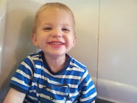 A young child with short blond hair smiles widely, showing his teeth. He is wearing a blue and white striped t-shirt and is sitting against a light-coloured wall, radiating happiness and joy.