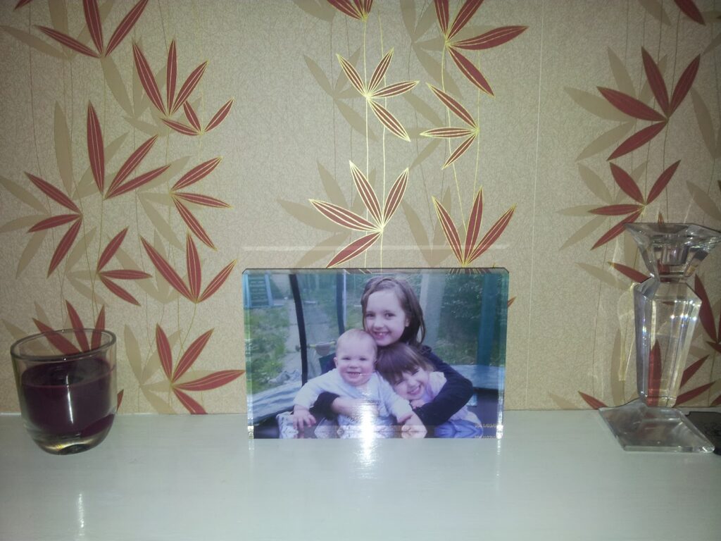 A framed photo of three smiling children sits on a white shelf. To the left is a small dark red candle in a glass holder. To the right is a clear glass decorative object. The background features beige wallpaper with a red leaf pattern.