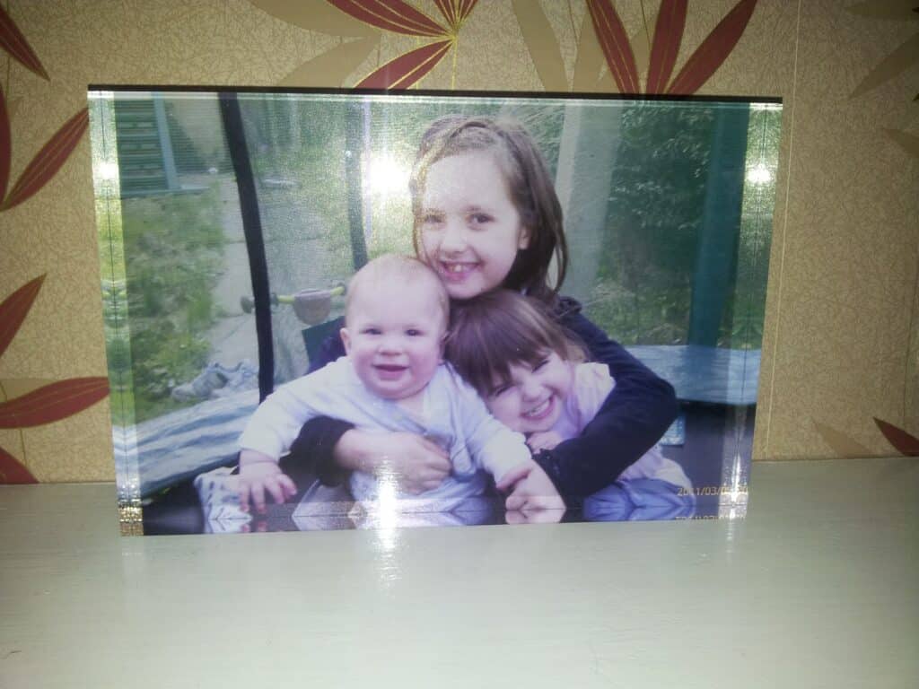 A framed photo of three smiling children, one older girl hugging a younger boy and another young girl. They are sitting close together outdoors, with greenery and a window in the blurred background. The frame rests on a light-coloured shelf.