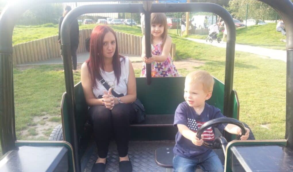 A woman with red hair sits in a playground vehicle, hands clasped, looking forward. A young girl stands behind her, smiling and holding onto a bar. A small boy with blond hair sits in front, pretending to drive, facing right. Grass and paths are visible outside.