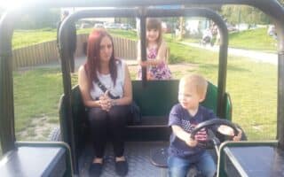 A woman with red hair sits in a playground vehicle, hands clasped, looking forward. A young girl stands behind her, smiling and holding onto a bar. A small boy with blond hair sits in front, pretending to drive, facing right. Grass and paths are visible outside.