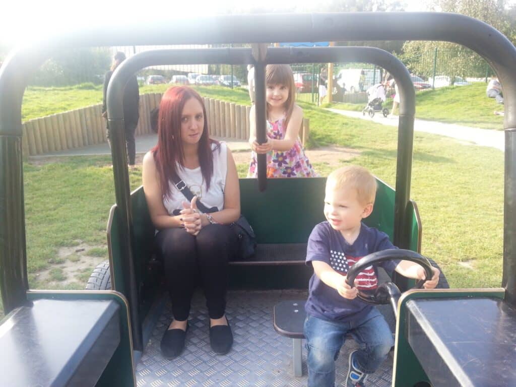 A woman with red hair sits inside a playground vehicle, hands clasped. A smiling young girl stands behind her, holding a bar, and a boy sits in front, gripping the steering wheel. Sunlight shines on the grassy park in the background.