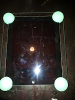 A tablet lies flat on a dark surface, with a greenish glow ball at each corner. The screen shows reflections of a bright light and surrounding objects, and appears smudged with fingerprints. Thin cords or strings outline the tablet’s rectangular shape.