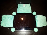 A tablet lies flat on a dark surface, surrounded at each corner by a light green ball and a folded soft green cloth. The tablet screen reflects a ceiling light above. There is a small tag on the top cloth. The arrangement appears intentional and symmetrical.