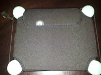 A black rectangular case with a flap covers a tablet. Four white rubber bumpers are attached to each corner of the tablet, slightly visible beyond the case. The device is on a dark surface, and a charging cable is visible at the top left.