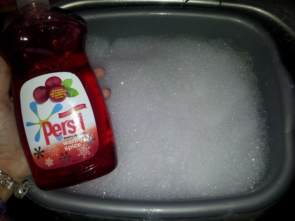A hand holds a red bottle of Persil washing-up liquid labelled "Warm Spice" next to a grey basin filled with white soapy bubbles, likely in a kitchen sink. The person's wrist wears a beaded bracelet.