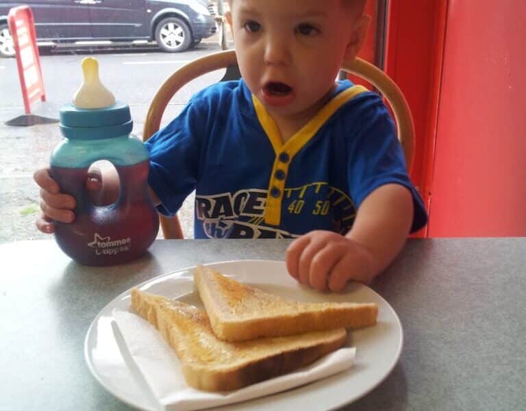 A young boy with short hair, wearing a blue and yellow shirt, sits at a table by a window. He holds a beaker and looks surprised, with his mouth open. In front of him is a white plate with two slices of toast on a serviette.