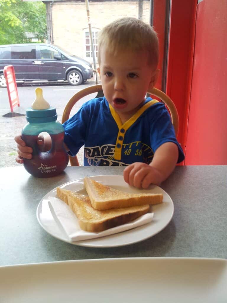 A young child with short blond hair sits at a table, holding a beaker with a blue lid in one hand and reaching for a plate of buttered toast in front of him. He wears a blue shirt and has a surprised or excited expression. A window and cars are visible outside.
