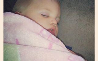 A young child with fair hair sleeps peacefully, wrapped snugly in a soft pink blanket with faint patterns, resting against a grey cushion. The child's face looks calm and serene, and the photo has a soft, cosy atmosphere.