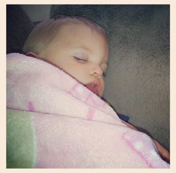 A young child with fair hair sleeps peacefully, wrapped snugly in a soft pink blanket with faint patterns, resting against a grey cushion. The child's face looks calm and serene, and the photo has a soft, cosy atmosphere.