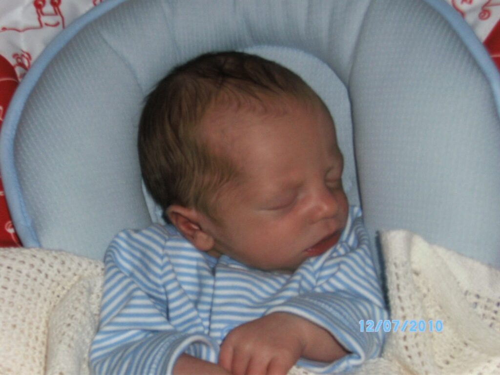 A newborn baby sleeps peacefully on a light blue cushioned surface, covered with a cream knitted blanket. The baby wears a blue and white striped babygrow, and the date “12/07/2010” is stamped in blue text on the lower right corner of the image.