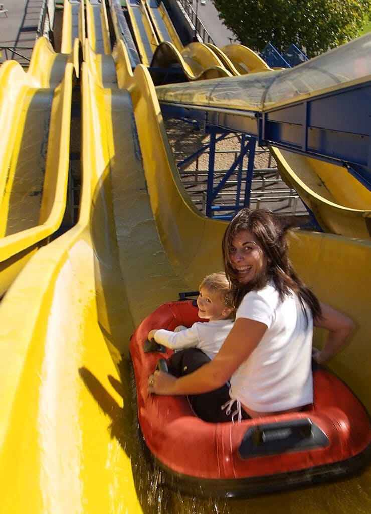 A woman and a young child sit together in a red inflatable raft on a yellow water slide at an amusement park. Both are smiling and facing backwards towards the camera as they begin their descent down the curving, bright slide.