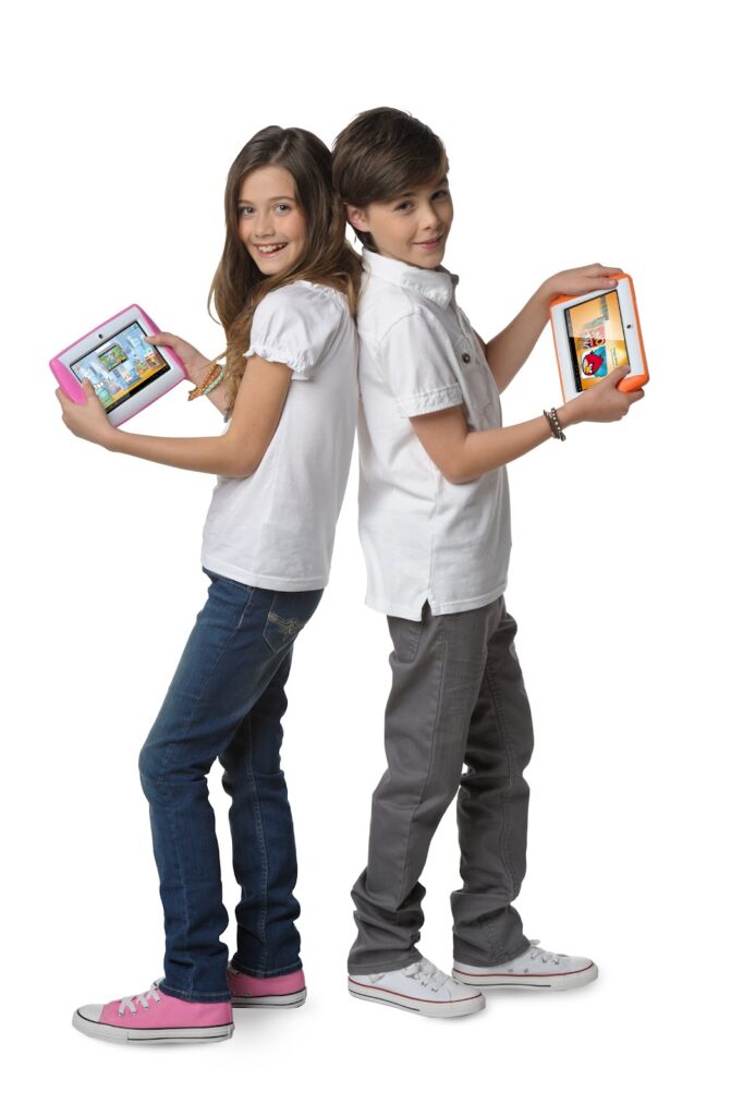 A girl and boy stand back-to-back, smiling, each holding a tablet. The girl wears a white shirt, jeans, and pink trainers; the boy wears a white shirt, grey trousers, and white trainers. Both tablets display colourful screens. The background is white.