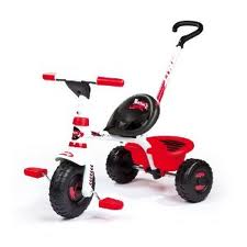 A red, white, and black tricycle with chunky wheels, a black seat, a tall rear handlebar for steering, and a small rear cargo area. The pedals are attached to the front wheel and the tricycle appears sturdy and designed for young children.