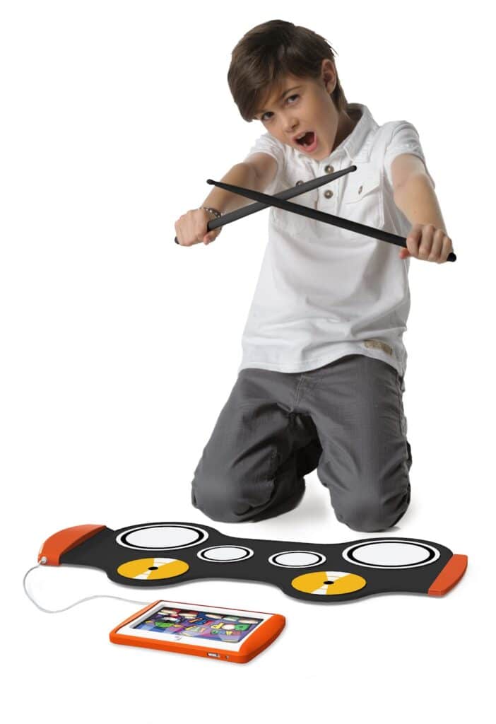 A boy kneels on the floor, enthusiastically holding two drumsticks above a black electronic drum pad placed in front of him. The pad is connected by a cable to a small orange tablet displaying a colourful interface.