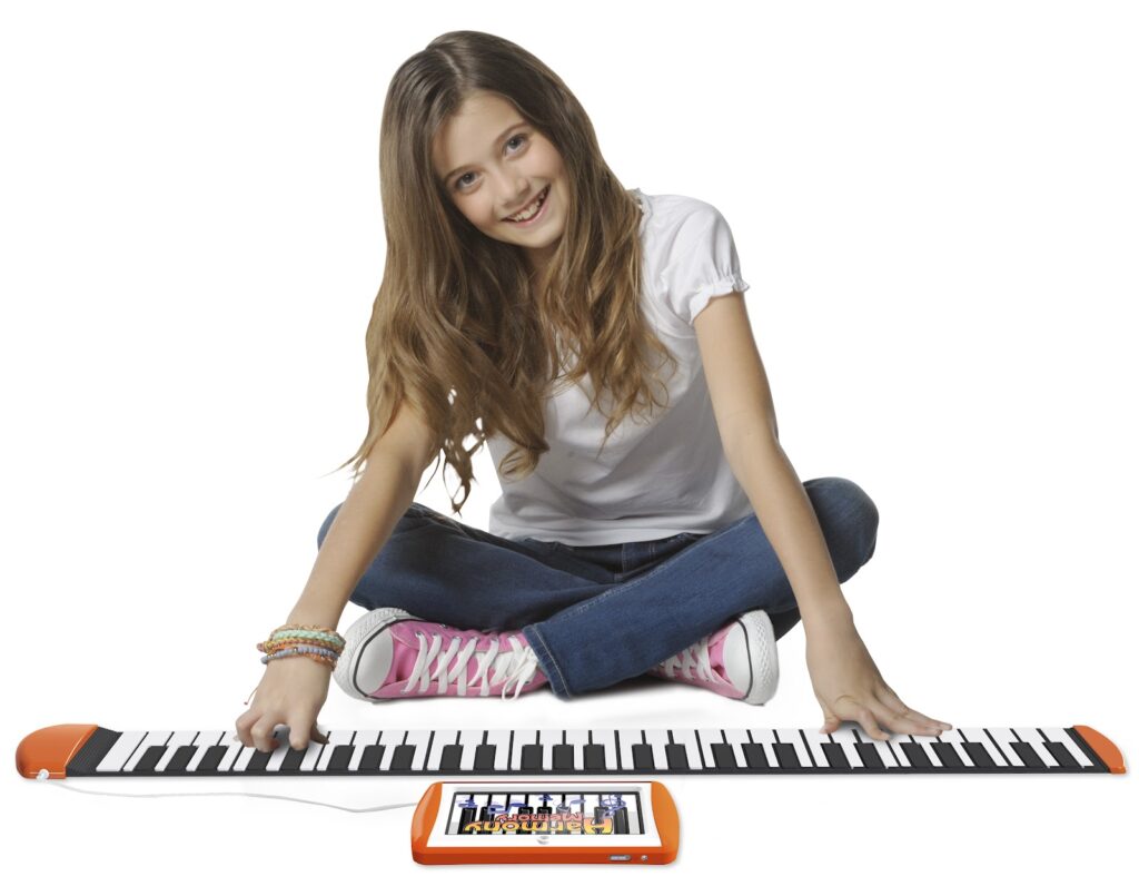 A smiling girl with long brown hair, wearing a white T-shirt, blue jeans, and pink trainers, sits cross-legged. She is playing a flexible roll-up piano keyboard laid out in front of her, with a small orange control panel attached.