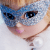 Close-up of a doll’s face wearing a sparkly blue mask over its eyes. The doll has blonde hair, glossy pink lips, and is dressed in a white fluffy outfit with a large pearl decoration near the collar. The overall feel is elegant and whimsical.