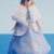 A blonde doll wearing a long, light blue ball gown with silver details and a white faux fur cape stands against a blue background. The doll has a tiara on her head and blue eyes, evoking a winter or princess theme.