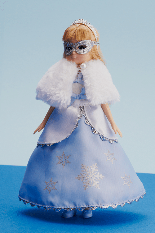 A blonde doll wears a silver tiara, silver mask, white faux fur cape, and a light blue gown with silver snowflake designs. She stands against a simple blue background. The outfit suggests a winter or princess theme.
