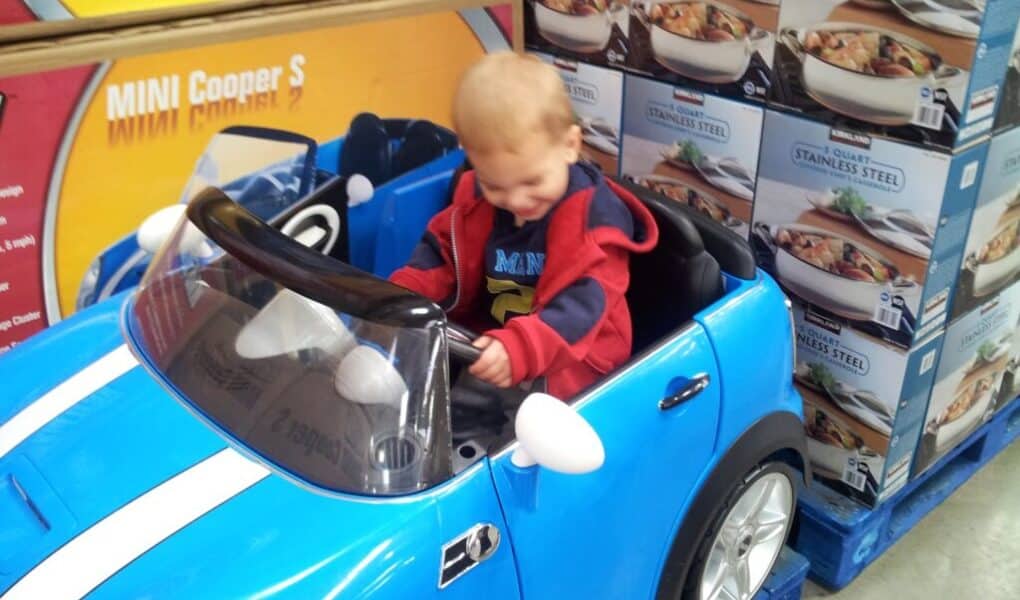A young child with short blond hair sits and smiles in a small blue toy Mini Cooper car. The car is indoors, next to stacked boxes of stainless steel cookware. The child wears a red and navy jacket and appears happy and playful.