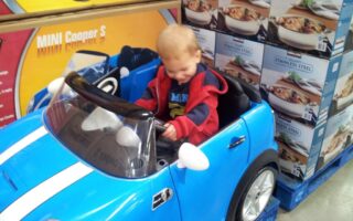A young child with short blond hair sits and smiles in a small blue toy Mini Cooper car. The car is indoors, next to stacked boxes of stainless steel cookware. The child wears a red and navy jacket and appears happy and playful.