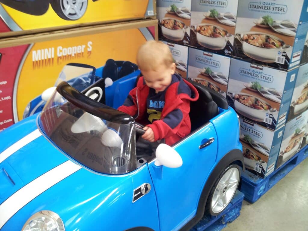 A young child with fair hair sits and smiles in a small blue toy Mini Cooper car at a shop. The car is beside a shelf stacked with boxes of stainless steel cookware. The child wears a red and blue jacket over a shirt.
