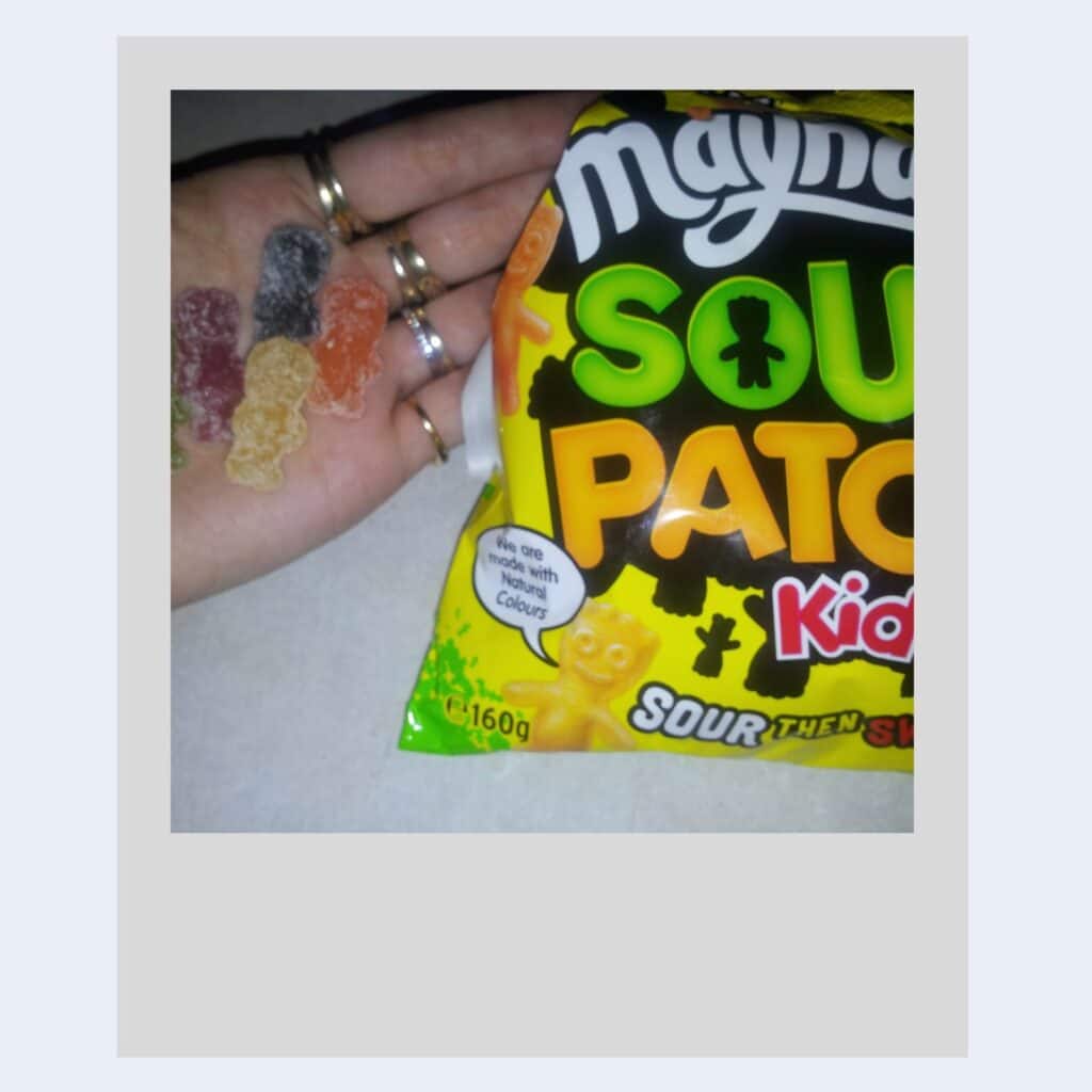 A hand with several rings holds five colourful Sour Patch Kids gummies next to a bright, partially opened 160g Maynards Sour Patch Kids sweet bag labelled "Sour Then Sweet" on a light surface.