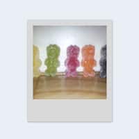 A photo shows five large gummy bear sweets in a row, each a different colour: yellow, green, pink, orange, and purple. They are placed upright on a wooden surface against a plain white background. The image has a white Polaroid-style border.
