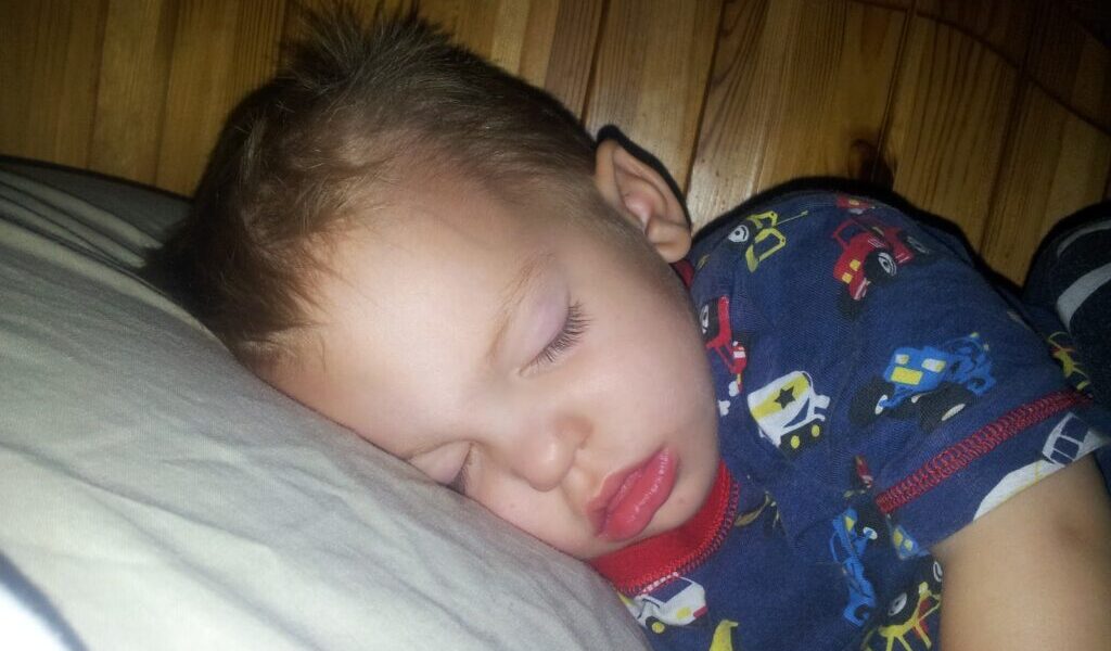 A young child with short light brown hair is sleeping on a white pillow, facing left. He is wearing dark blue pyjamas with colourful cars and lorries. The background shows a wooden headboard, and the lighting is soft, giving a peaceful atmosphere.