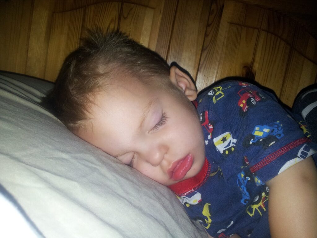 A young child with short light brown hair is sleeping on a white pillow, facing left. He is wearing a dark blue shirt with colourful cars printed on it, and his lips are slightly parted. The background is a wooden headboard.