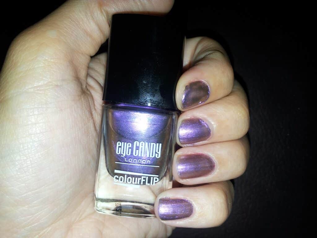 A hand with short nails painted metallic purple holds a bottle of Eye Candy London colourFLIP nail varnish. The varnish bottle has a black cap and clear label, and the background is dark, making the nail colour stand out.