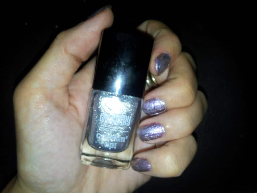 A hand with shimmery purple nail varnish holds a bottle of glittery grey nail varnish with a black cap, against a dark background. The nails are neatly painted and reflect light, highlighting the sparkly finish.