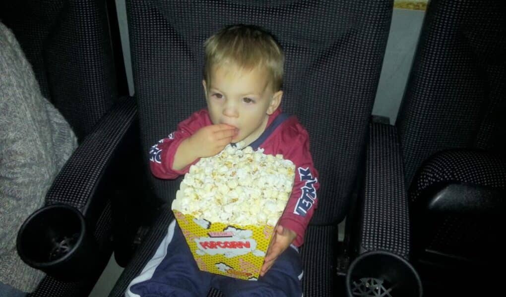 A young child sits in a cinema seat, holding a large box of popcorn on their lap and eating with one hand. The child wears a red jacket, blue trousers, and white socks. Armrests and cup holders are visible on both sides of the seat.