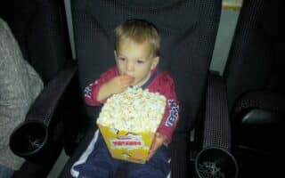 A young child sits in a cinema seat, holding a large box of popcorn on their lap and eating with one hand. The child wears a red jacket, blue trousers, and white socks. Armrests and cup holders are visible on both sides of the seat.