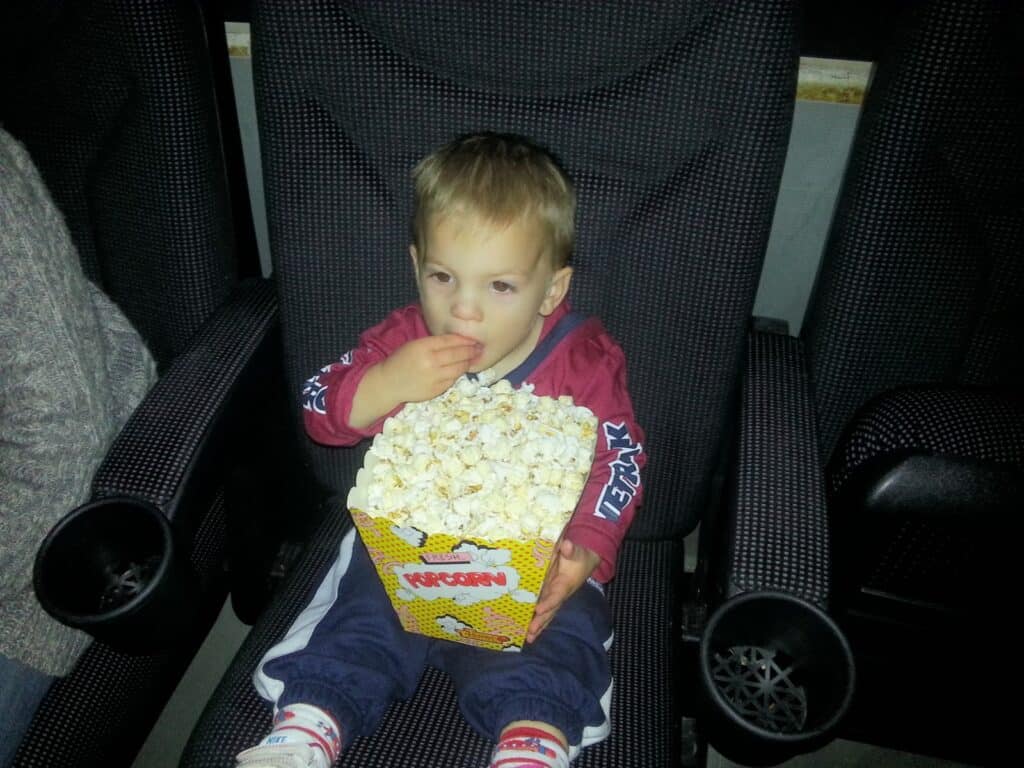 A young child sits in a large cinema seat, wearing a red jumper and blue trousers, holding a big box of popcorn on their lap and eating some. The child looks focused, and the seat’s dark armrests and cupholders are visible.