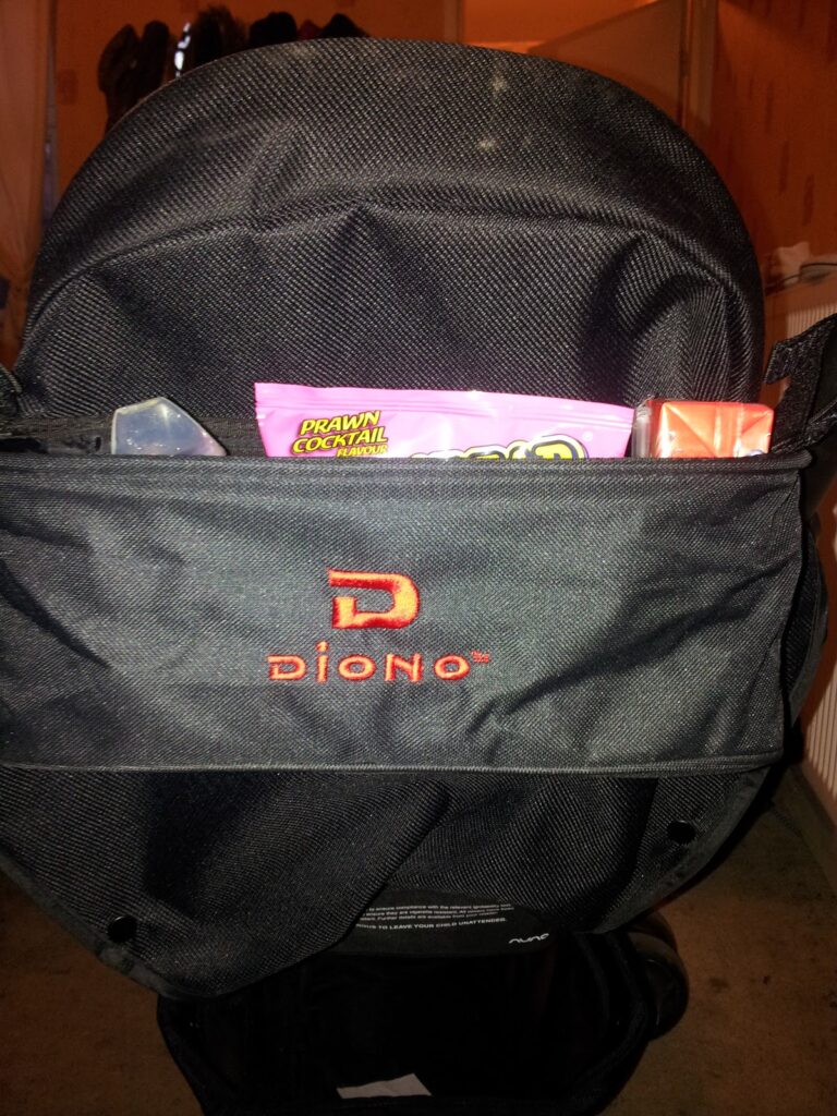 A black Diono pushchair pocket with red logo contains a pink pack of prawn cocktail-flavoured crisps and a rectangular orange carton, all situated indoors on a carpeted floor with a radiator and shoes visible in the background.