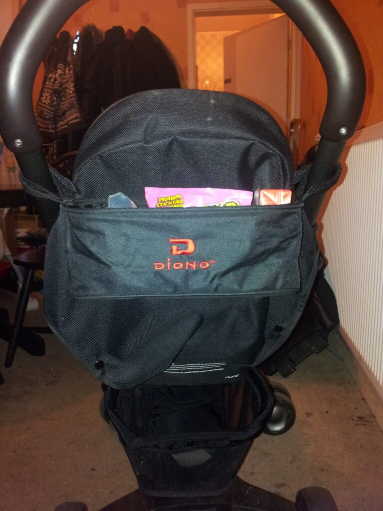 A black Diono pushchair seen from the back, with a storage pouch holding a pink snack packet and an orange item. The pushchair is indoors on a carpeted floor, and jackets hang on a rack in the background with a white radiator on the right.