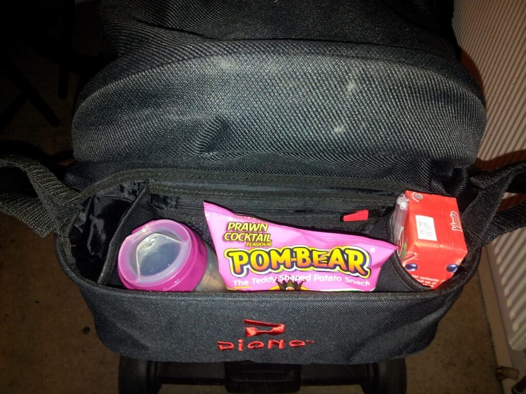 An open black Diono pushchair bag contains a pink-lidded baby bottle, a pink bag of POM-BEAR prawn cocktail crisps, and a small red carton with white text. The bag is open, showing the items neatly placed inside.