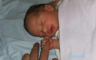 A newborn baby sleeps on a light blue blanket, wrapped in a pale purple cloth. The baby gently grasps an adult’s finger with one hand. The baby has dark hair and a peaceful expression. A timestamp in blue text shows the date: 03/07/2010.