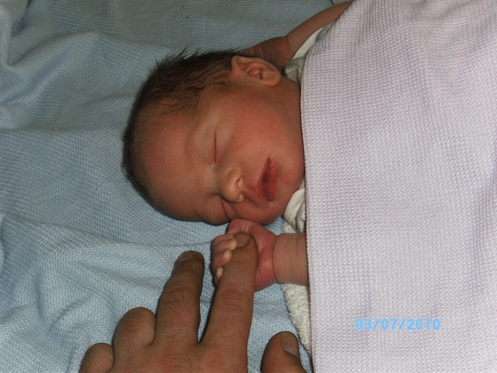 A newborn baby sleeps peacefully on a light blue blanket, wrapped in a pale lavender cloth. The baby gently grasps an adult's finger with its tiny hand. The date “03/07/2010” is visible in blue text in the lower right corner.