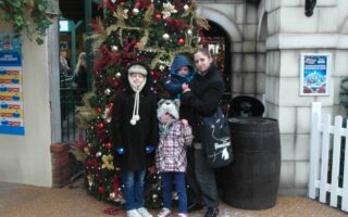 A woman and three children in winter coats pose in front of a decorated Christmas tree outside a building. The children wear hats and the youngest is held in the woman's arms. The scene has festive baubles and a wooden barrel beside a white picket fence.