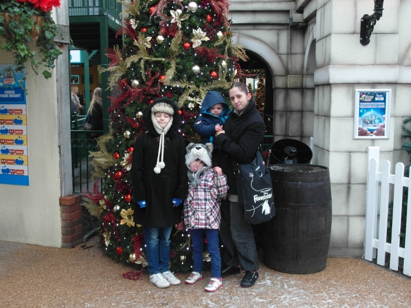 A woman and three children in winter coats pose in front of a decorated Christmas tree outside a building. The children wear hats and the youngest is held in the woman's arms. The scene has festive baubles and a wooden barrel beside a white picket fence.