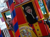 Two people wearing winter hats smile from the window of a brightly coloured carnival train, decorated with stars and a clown illustration. The train has red, yellow, and blue stripes, and a carnival atmosphere is visible in the background.