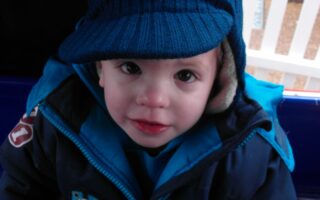 A young child with big brown eyes and rosy cheeks looks up at the camera, wearing a blue jacket with a Thomas the Tank Engine badge and a blue knitted hat with ear flaps. The background shows a white fence and some outdoor flooring.
