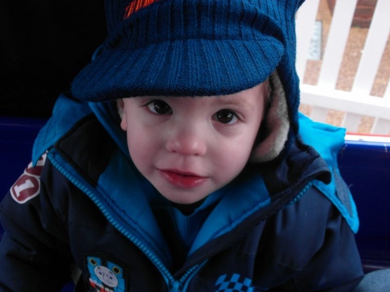 A young child with big brown eyes and rosy cheeks looks up at the camera, wearing a blue jacket with a Thomas the Tank Engine badge and a blue knitted hat with ear flaps. The background shows a white fence and some outdoor flooring.