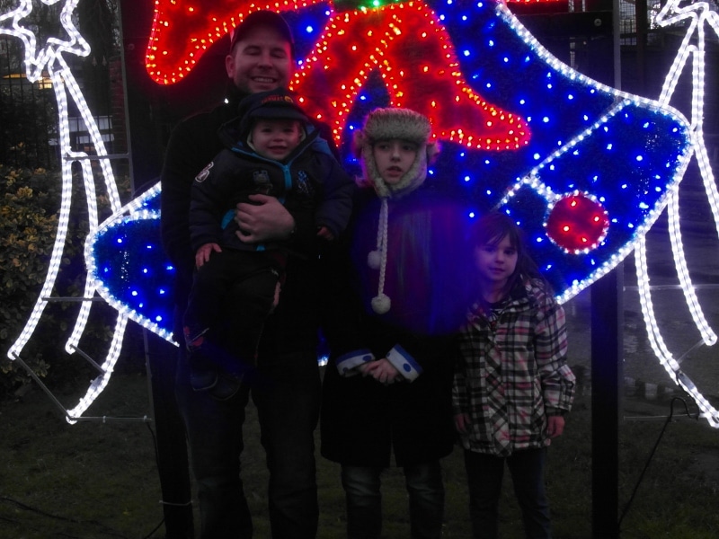 A man holding a small child stands beside two children in winter coats and hats. They are outdoors at night in front of a large, brightly lit holiday display shaped like a bell with red, blue, and white lights. All four look towards the camera.