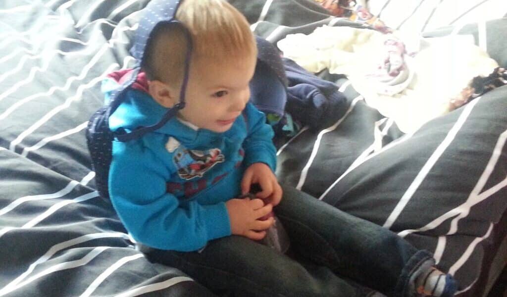 A toddler sits on a bed with grey and white striped bedding, smiling and wearing a blue hoodie. A blue bag or hat is draped over the child’s head. Clothes are scattered on the bed in the background. The scene is brightly lit.