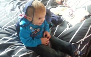 A toddler sits on a bed with grey and white striped bedding, smiling and wearing a blue hoodie. A blue bag or hat is draped over the child’s head. Clothes are scattered on the bed in the background. The scene is brightly lit.