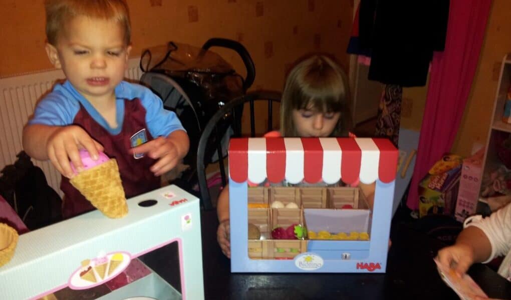 A young boy holds a toy ice cream cone beside a toy ice cream set, while a young girl plays with a pretend fruit stall. Another person’s hand appears on the right, possibly handling toy money. The setting appears to be a playroom or dining area.